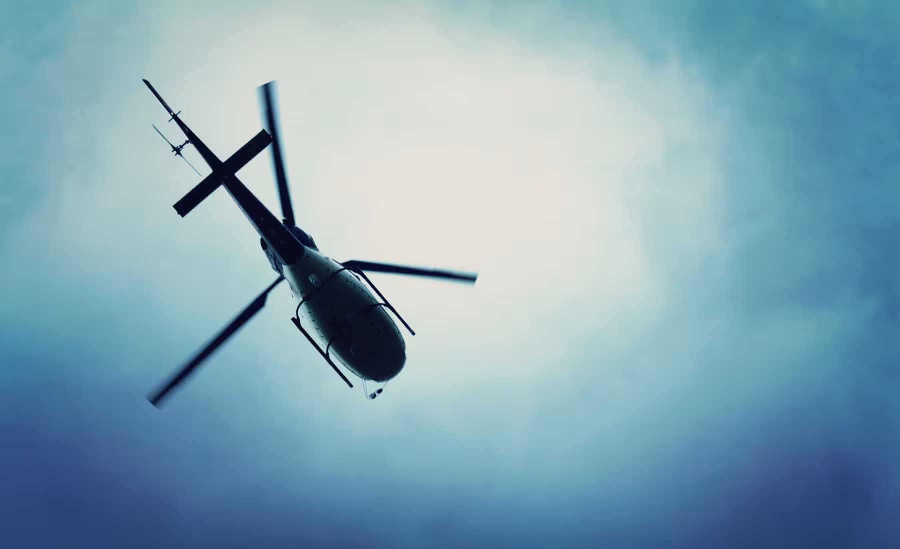 A helicopter viewed from below, flying against a cloudy sky with a blue gradient. The rotor blades appear slightly blurred, emphasizing motion.