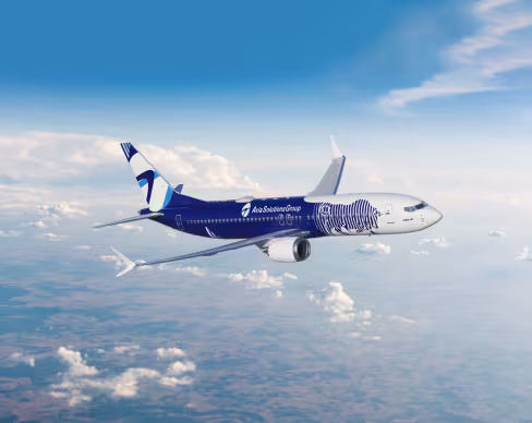 A Boeing 737 in Avia Solutions Group livery, featuring a dark blue fuselage with a white-blue zebra design. The aircraft flies against a bright sky with scattered clouds and landscape below.