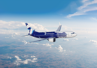 A Boeing 737 in Avia Solutions Group livery, featuring a dark blue fuselage with a white-blue zebra design. The aircraft flies against a bright sky with scattered clouds and landscape below.