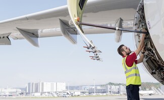 A SWISS line maintenance technician performing maintenance on an aircraft engine at the airport, with the engine panel open and airport facilities visible in the background.