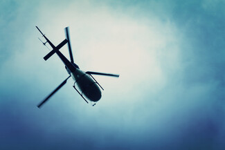 A helicopter viewed from below, flying against a cloudy sky with a blue gradient. The rotor blades appear slightly blurred, emphasizing motion.