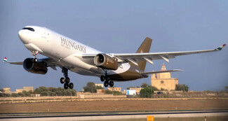 A Hungary-registered Airbus A330 takes off, with landing gear extended. The white fuselage features "HUNGARY" and gold-brown accents. The Hungarian flag colors appear on the winglets, with a historical building in the background.