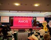 A man stands on a stage with a red screen behind him displaying the word "AMOS." People are seated at tables in front of the stage, with flower arrangements on either side.